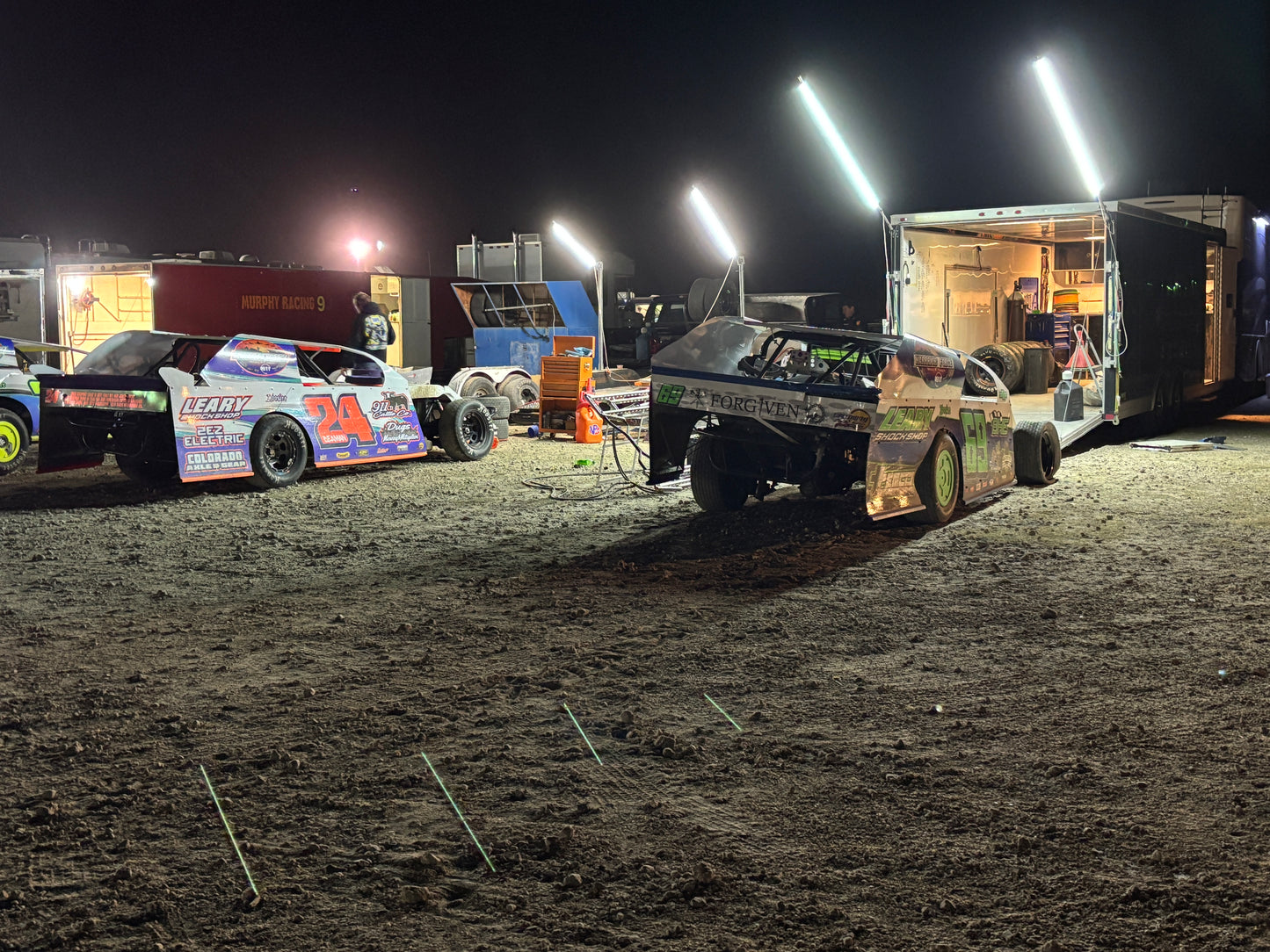 Two race cars on a dirt track at night with bright lights illuminating the scene.