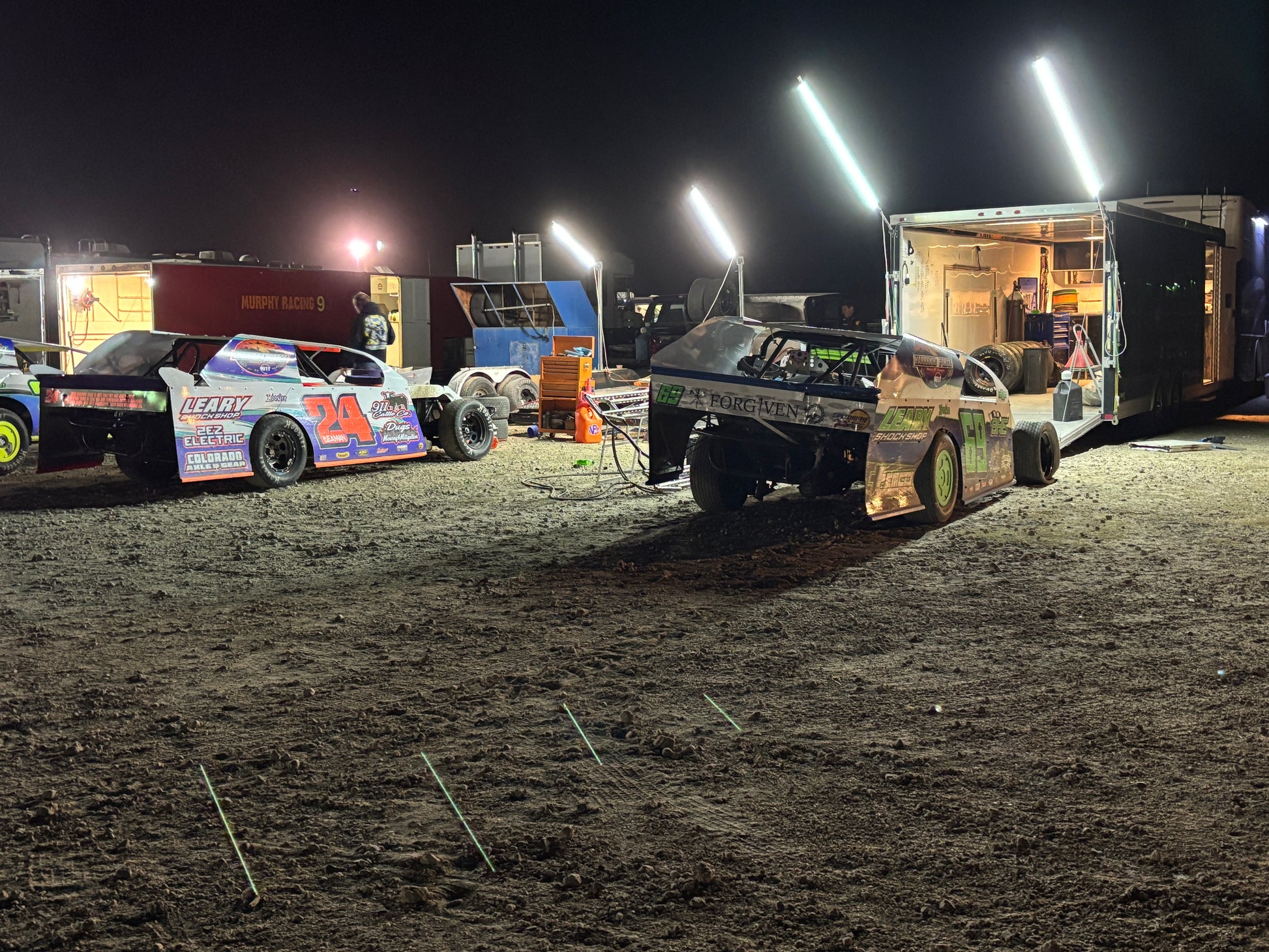 Two race cars on a dirt track at night with bright lights illuminating the scene.